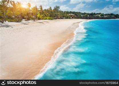 Aerial view of white sandy beach, ocean with waves, umbrellas, green palms at sunset. Summer holiday in Zanzibar, Africa. Tropical landscape with palm trees, sand, blue water, people, sky. Top view