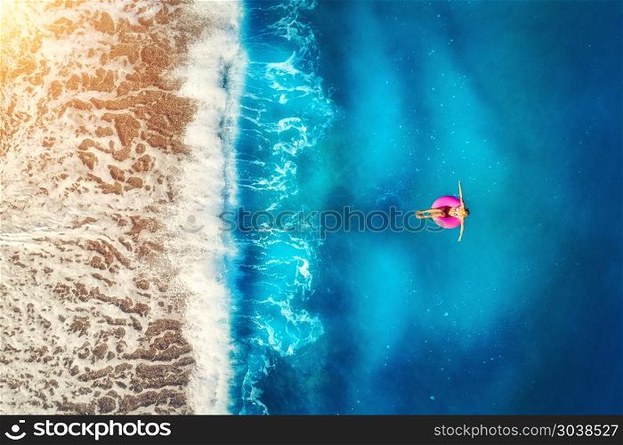 Aerial view of woman swimming on the pink swim ring in the transparent sea and beautiful waves in Europe. Summer landscape with girl, beach, blue water at sunset. Top view. Travel and holiday. Resort. Aerial view of woman swimming on the pink swim ring in the sea