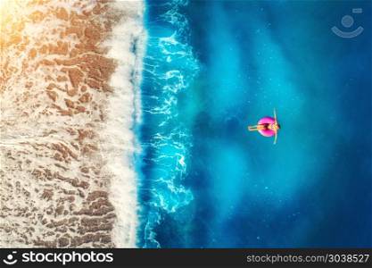 Aerial view of woman swimming on the pink swim ring in the transparent sea and beautiful waves in Europe. Summer landscape with girl, beach, blue water at sunset. Top view. Travel and holiday. Resort. Aerial view of woman swimming on the pink swim ring in the sea