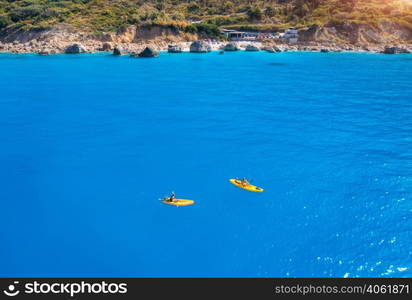 Aerial view of yellow kayaks in blue sea at sunset in summer. Man and woman on floating canoe in clear azure water. Lefkada island, Greece. Tropical landscape. Sup boards. Active travel. Top view