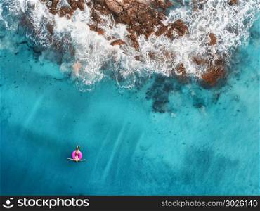 Aerial view of young woman swimming on the pink swim ring in the transparent turquoise sea in Maldives. Summer seascape with girl, beach, beautiful waves, rocks, blue water at sunset. Top view. Nature