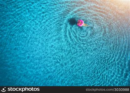 Aerial view of young woman swimming on the pink swim ring in the transparent blue sea at sunrise. Summer aerial seascape with girl, clear water in the morning. Top view. Travel and holiday. Resort. Aerial view of young woman swimming in the sea with transparent water