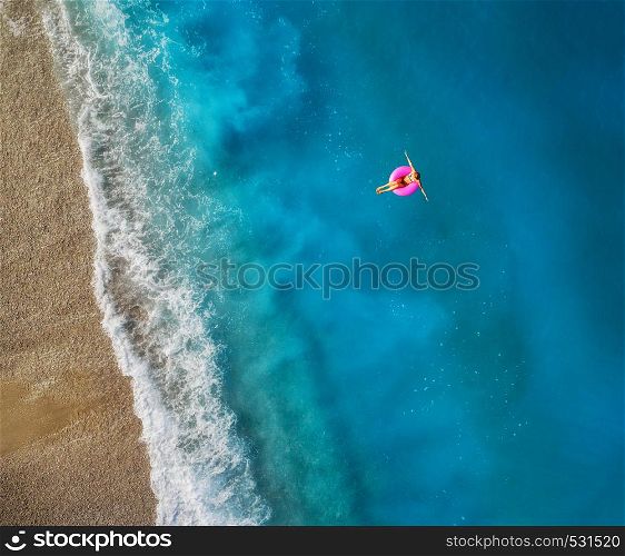 Aerial view of young woman swimming on the pink swim ring in the transparent turquoise sea in Oludeniz,Turkey. Summer seascape with girl, beautiful waves, blue water in sunny day. Top view from drone. Aerial view of young woman swimming on the pink swim ring