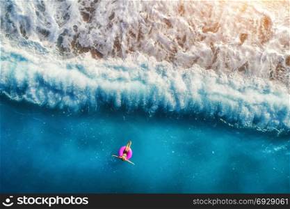 Aerial view of young woman swimming on the pink swim ring in the transparent turquoise sea in Oludeniz. Summer seascape with girl, beach, beautiful waves, blue water at sunset. Top view from drone. Aerial view of young woman swimming in the sea
