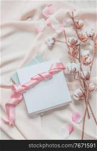 Aesthetic lifestyle with notebook, white box and pink ribbon on beige fabric background with cotton branches and pink petals. Top view. Beauty