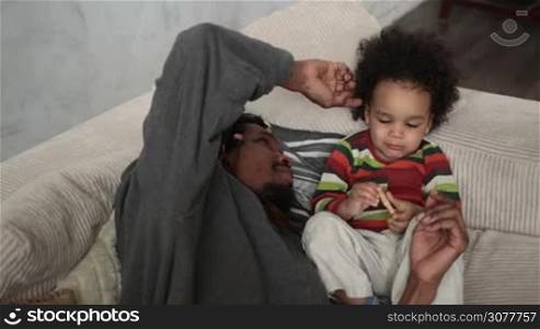 Affectionate african american father with dreadlocks caressing curly mixed race toddler son&acute;s hair while lying on the sofa. Loving dad and little child spending time together, father tenderly stroking son&acute;s hair. Slow motion.