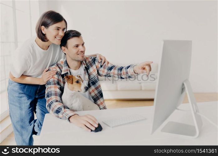 Affectionate brunette wife supports husband with doing project work, happy man points into monitor, tells something, shares his ideas, works together with pedigree dog, pose in spacious cabinet