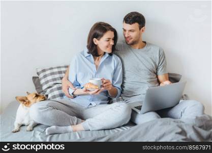 Affectionate couple in love embrace in bedroom, warch film online on laptop computer, have breakfast, enjoy domestic atmopshere and their favourite pet lies in bed. People, relatioships concept