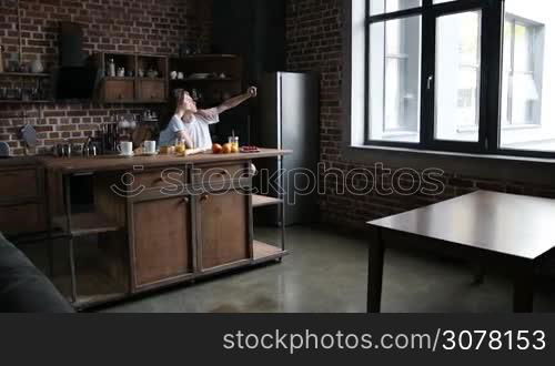 Affectionate couple sitting at the kitchen table and making selfie with smart phone while having breakfast in the morning. Beautiful couple relaxing, enjoying good time, fooling around while taking self-portrait on mobile phone at home. Slow motion.