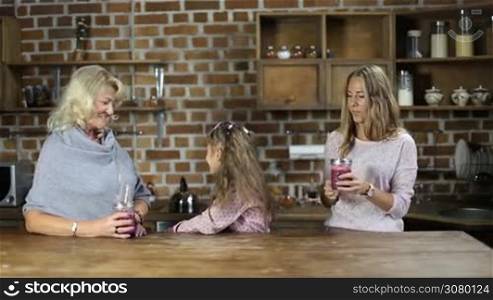 Affectionate daughter embracing and kissing her pretty mother while multi generation family having healthy breakfast in the kitchen. Happy grandmother, daughter and lovely granddaughter drinking fresh berry smoothies while spending leisure at home.