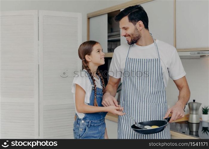 Affectionate father in apton fries eggs, teaches small daughter how to cook, pose together at kitchen, prepare delicious breakfast, look at each other with smile. Children, fatherhood, domestic life