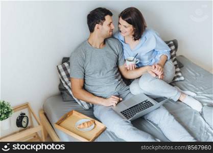 Affectionate female and male look at each other with great love, enjoy togetherness in bedroom, have good relationships, use laptop computer for entertainment, drink tea with croissant