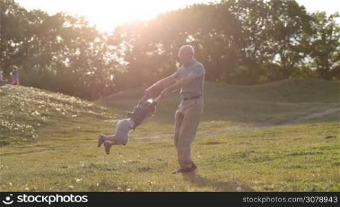 Affectionate grandad playing and spinning his adorable grandson around while family enjoying leisure in park in glow of amazing sunset. Handsome grandpa in eyeglasses having fun with grandchild, spinning his laughing kid in circle outdoors.