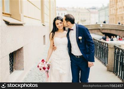 Affectionate handsome bridegroom with beard kisses his future wife. Lovely brunette bride wears nice dress and holds bouquete, has walk with lover on bridge. Marriage and celebration concept