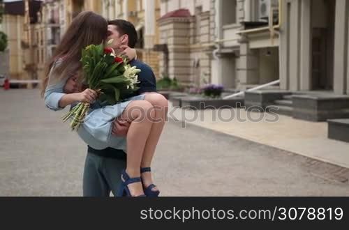 Affectionate hipster man holding his charming girlfriend in his arms in city street background. Joyful young woman with bunch of flowers embracing man&acute;s neck gently and kissing her boyfriend while young teenager man carrying his cute girl in his arms