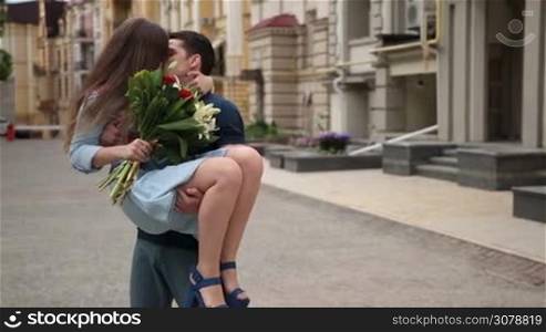 Affectionate hipster man holding his charming girlfriend in his arms in city street background. Joyful young woman with bunch of flowers embracing man&acute;s neck gently and kissing her boyfriend while young teenager man carrying his cute girl in his arms