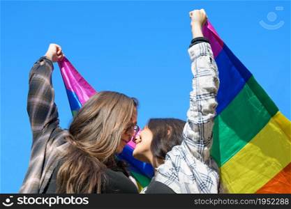 Affectionate lesbian couple holding an LGBT flag under blue sky. High quality photo.. Affectionate lesbian couple holding an LGBT flag under blue sky.
