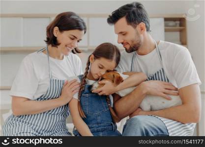Affectionate little girl embraces pedigree dog with love, mother and father pose near, spend free time at kitchen, prepare delicious breakfast. Happy family and their pet love each other very much