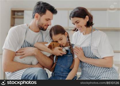 Affectionate little girl embraces pedigree dog with love, mother and father pose near, spend free time at kitchen, prepare delicious breakfast. Happy family and their pet love each other very much