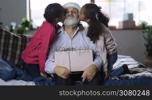 Affectionate little mixed race granddaughters kissing excited grandfather with gift on his lap on the cheeks and embracing at christmas. Little girls giving a kiss to grandpa in santa hat while happy family spending time together on winter holidays.