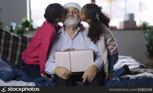 Affectionate little mixed race granddaughters kissing excited grandfather with gift on his lap on the cheeks and embracing at christmas. Little girls giving a kiss to grandpa in santa hat while happy family spending time together on winter holidays.