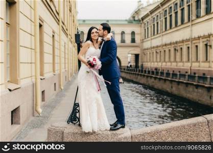 Affectionate married couple kiss passionately as stand on bridge near river in ancient city, celebrate their wedding, being happy to creat their friendly family. Romantic newlyweds pose outdoors