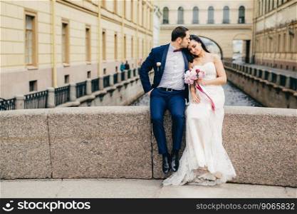 Affectionate married couple sit on bridge togetherm enjoy fresh air, have good relationship, kiss each other. Young female in white wedding dress recieves kiss from husband. Togetherness concept