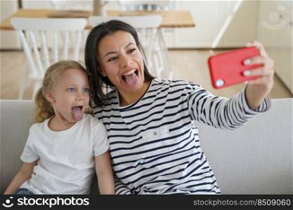 Affectionate mother and kid are taking selfie. People having fun, playing and show tongues. Young european mum makes photo of happy moment with little daughter. Cheerful family has leisure at home.. Affectionate mother and kid are taking selfie. Young european mum makes photo with little daughter.