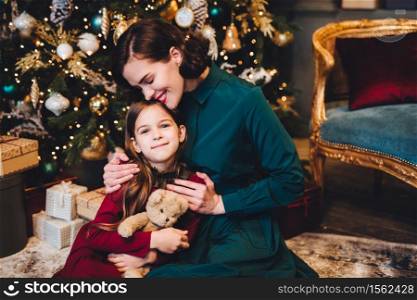 Affectionate mother embraces her little daughter as sit together near decorated Christmas tree. Small adorable girl being glad to recieve New Year present from her mom. Relationship concept.