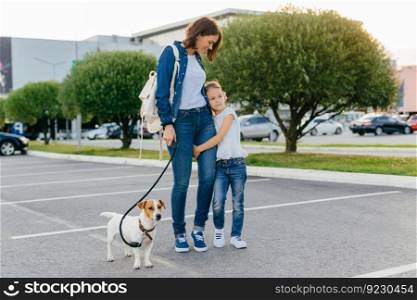Affectionate mother embraces small daughter, have outdoor stroll together with their favourite pet, enjoy good day and weather, pose against city background. Family, urban style and day off concept