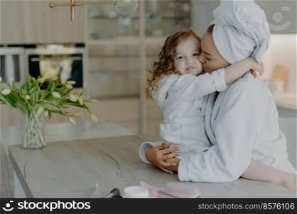 Affectionate mother wearing dressing gown wrapped bath towel on head embraces and kisses with love her small daughter pose together at home against cozy interior feel refreshed after taking shower