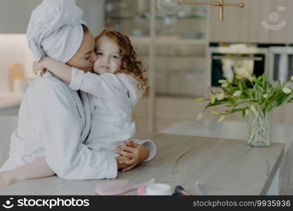 Affectionate mother wearing dressing gown wrapped bath towel on head embraces and kisses with love her small daughter pose together at home against cozy interior feel refreshed after taking shower