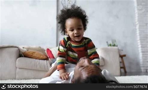 Affectionate playful african american father with dreadlocks kissing his cute little mixed race child while lying on his back on the floor at home. Young joyful dad with lovely toddler boy lying on his chest having fun and playing in living room.