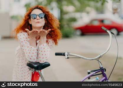 Affectionate redhead woman sends air kiss to lover on street, keeps palms near mouth, wears round shades, dress, leans at bicycle, has active rest outdoor, poses on street. Recreational time concept