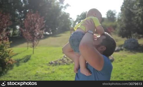 Affectionate young father with beard raising his laughing infant son up in the air and kissing his belly in the park on sunny summer day. Father playing with smiling baby boy outdoors. Slow motion. Steadicam stabilized shot.