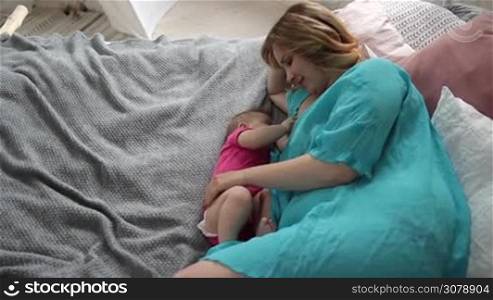 Affectionate young mom stroking her baby girl while lying on bed and breastfeeding her newborn child. Top angle view. Sweet infant baby drinking breastmilk. Slow motion. Steadicam stabilized shot.