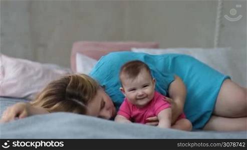 Affectionate young mother kissing her infant daughter while lying in bed in the morning. Happy beautiful mom kissing and hugging her adorable baby girl while relaxing in bed at home. Family enjoying time together.