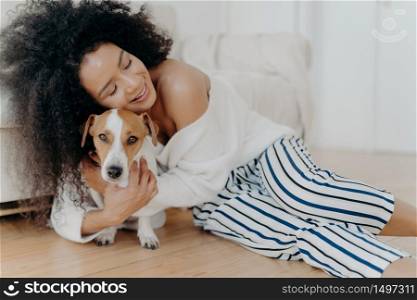 Affectionate young woman hugs dog with love and care, keeps eyes closed from pleasure, smiles gently, has healthy dark skin, poses on floor, petting animal. People, friendship and pets concept
