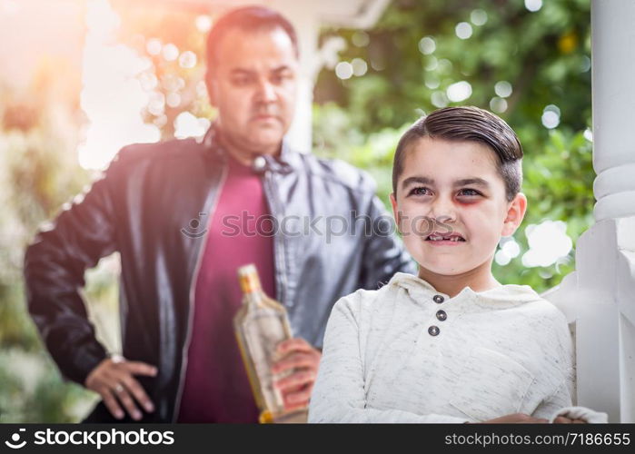 Afraid and Bruised Mixed Race Boy In Front of Angry Man Holding Bottle of Alcohol.