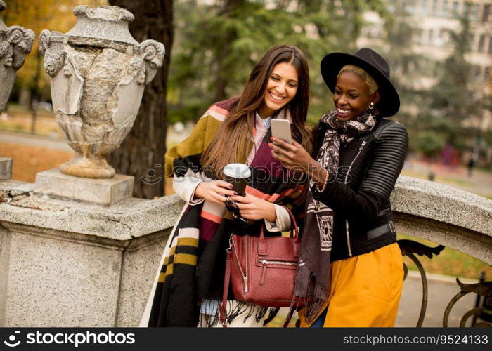 African american and caucasian woman posing outside with mobile phone and a cup of coffee to go in autumn