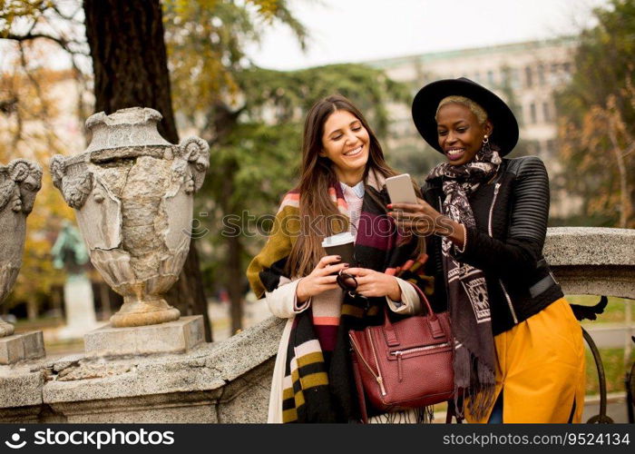 African american and caucasian woman posing outside with mobile phone and a cup of coffee to go in autumn