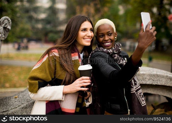African american and caucasian woman posing outside with mobile phone and a cup of coffee to go in autumn