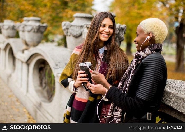 African american and caucasian woman posing outside with mobile phone and a cup of coffee to go in autumn