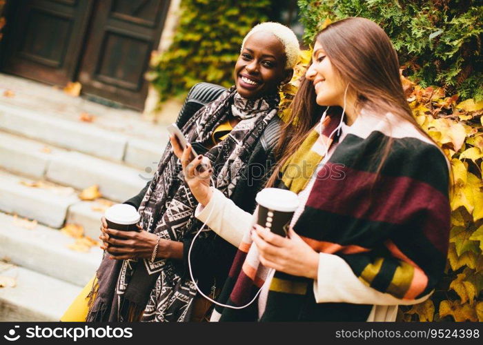 African american and caucasian woman posing outside with mobile phone and a cup of coffee to go in autumn