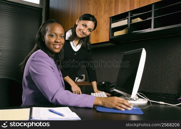 African-American and Indian young adult business women working at computer in office smiling and looking at viewer.