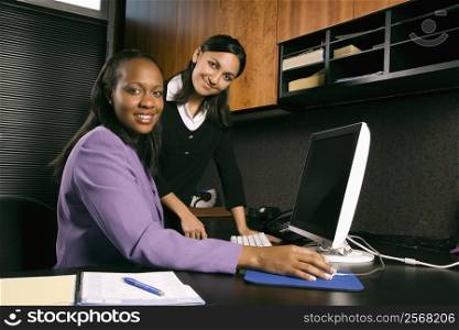African-American and Indian young adult business women working at computer in office smiling and looking at viewer.