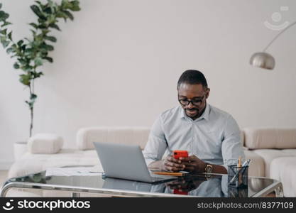 African american businessman looking at smartphone screen, sitting at laptop, working on online project, financial statistics. Black businessperson messaging with business partner by phone at office.. African american businessman uses phone app messages with business partner at office. E-business