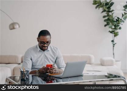 African american businessman looking at smartphone screen, sitting at laptop, working on online project, financial statistics. Black businessperson messaging with business partner by phone at office.. African american businessman uses phone app messages with business partner at office. E-business