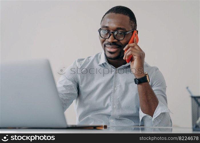 African american businessman makes business call, talking on phone, discussing working at laptop. Smiling black male manager in glasses holding smartphone, consulting client, sitting at office desk.. African american businessman makes business call, talking on phone, discussing working at laptop