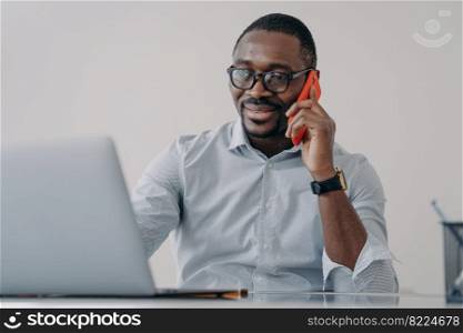 African american businessman makes business call, talking on phone, discussing working at laptop. Smiling black male manager in glasses holding smartphone, consulting client, sitting at office desk.. African american businessman makes business call, talking on phone, discussing working at laptop